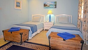 A bedroom with two beds, a nightstand with a lamp, a wooden chest, a patterned rug, and two framed pictures on the wall. - Arcadia by Blue Sky Luxury (Worthing)