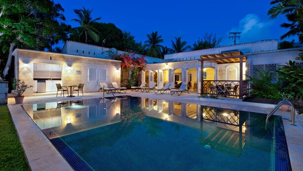 Una piscina con sillas de descanso y un patio cubierto, rodeada de abundante vegetación y palmeras.