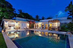 A swimming pool with lounge chairs and a covered patio area, surrounded by lush greenery and palm trees. - Todmorden by Blue Sky Luxury (Gibbes)