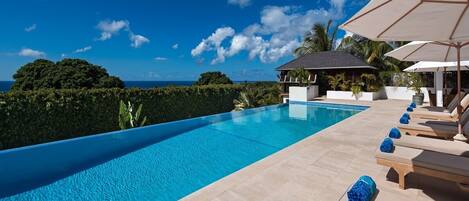 A swimming pool with lounge chairs and umbrellas, surrounded by greenery and a view of the ocean.