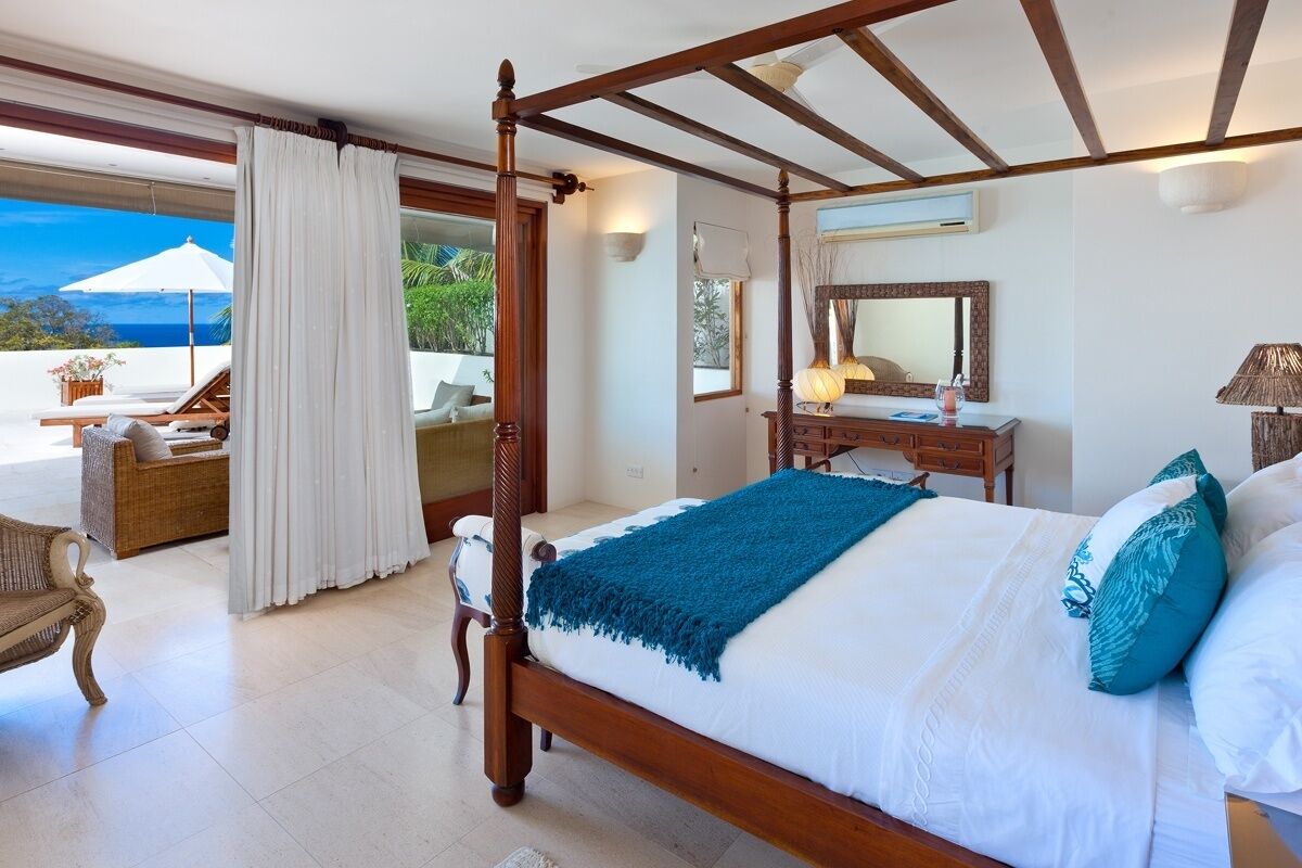 A four-poster bed with a blue throw, a wooden nightstand, a mirror, and a view of the ocean.