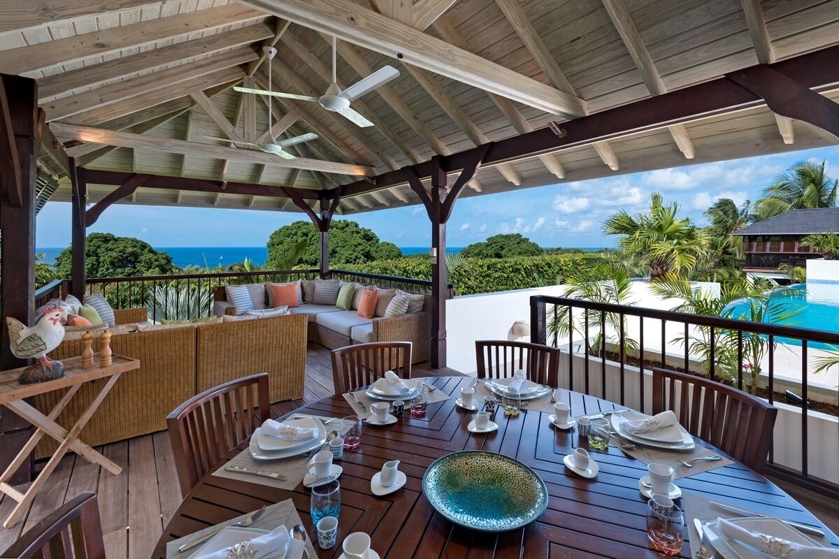 A covered outdoor dining area with a view of the ocean, wooden furniture, and a table set for a meal.