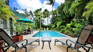 A pool area with lounge chairs, a table, and a building with a green awning. - Jessamine by Blue Sky Luxury (Gibbes)