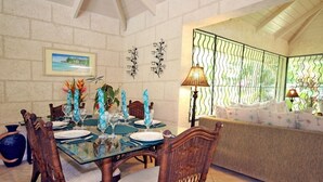 A dining area with a glass table, wicker chairs, and a sofa with a patterned cushion. - Jessamine by Blue Sky Luxury (Gibbes)