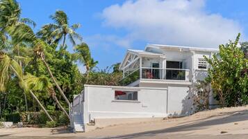 A white beachfront house with a balcony, surrounded by palm trees and a clear sky.