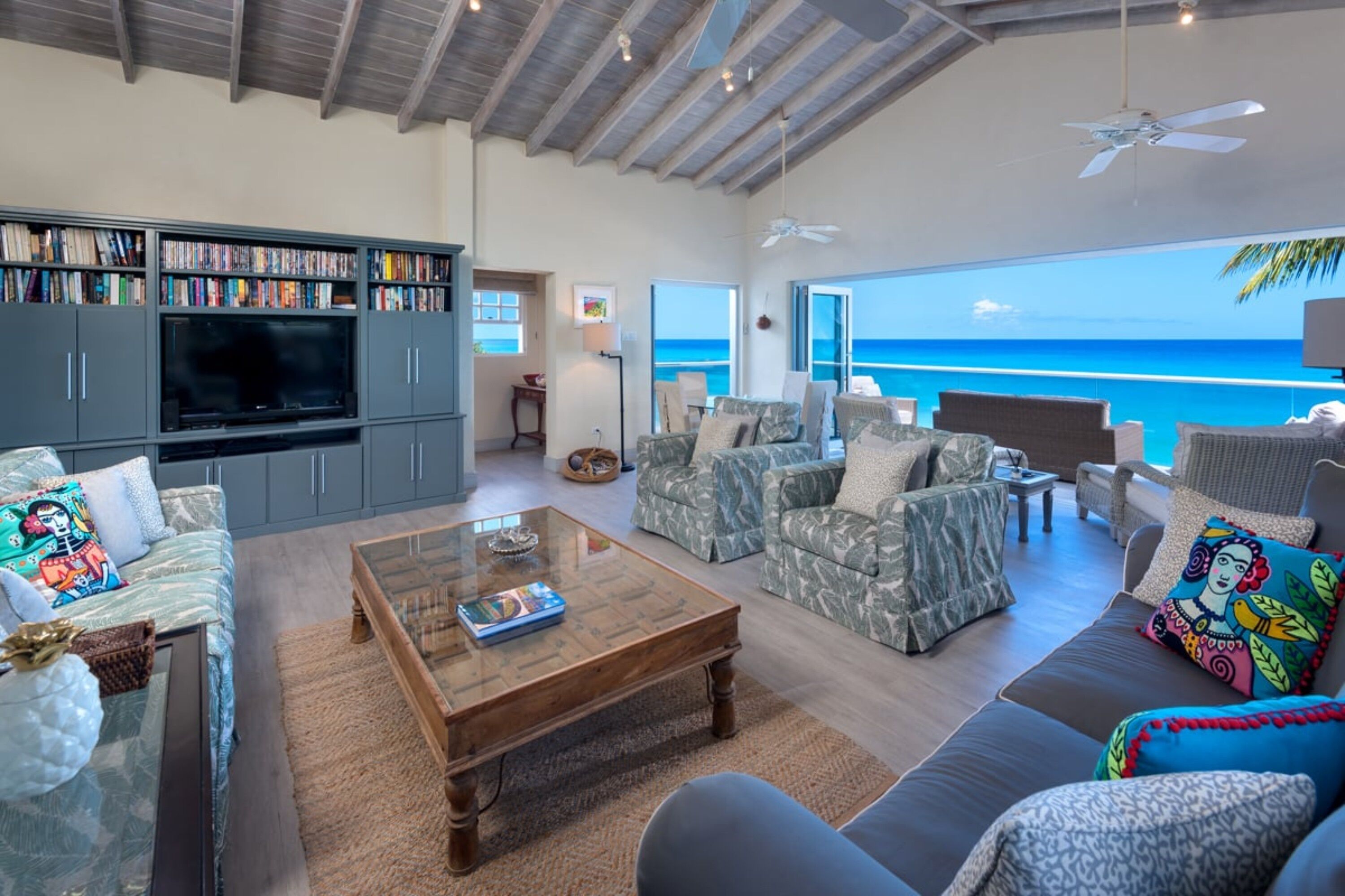 A spacious living room with a large flat-screen TV, a bookshelf, and a view of the ocean.