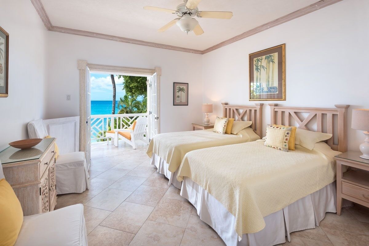 A hotel room with two beds, a ceiling fan, a balcony with a view of the ocean, and a door leading outside.