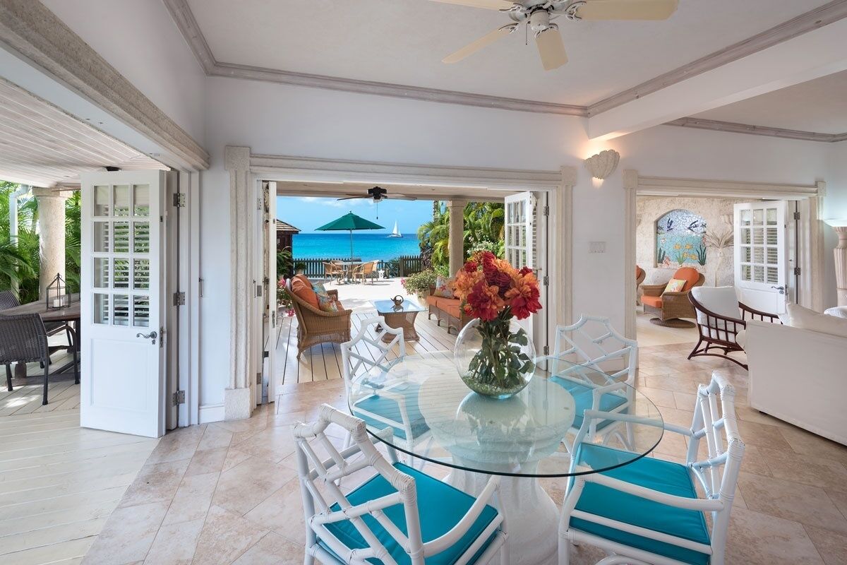 A spacious room with a glass table, white wicker chairs, a glass vase with flowers, and a view of the ocean.