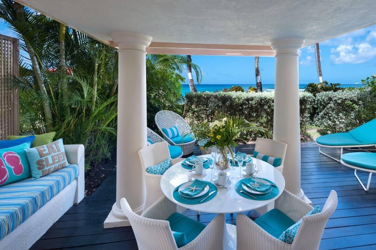 A covered outdoor dining area with white wicker furniture and blue cushions, overlooking a beach.