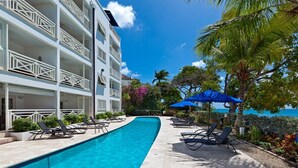 A swimming pool with lounge chairs and umbrellas, a multi-story building with balconies, and a view of the sea. - Waterside 402 by Blue Sky Luxury (Paynes Bay)