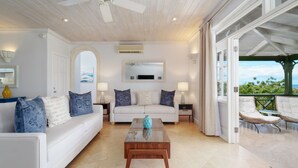 Een ruime woonkamer met een witte bank, blauwe kussens, een houten salontafel en uitzicht op het buitenleven door grote glazen deuren.