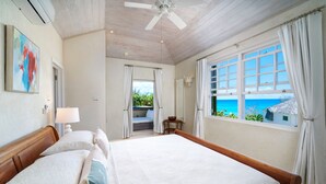 Een groot bed met witte lakens, een plafondventilator en uitzicht op de oceaan door een raam met witte gordijnen.