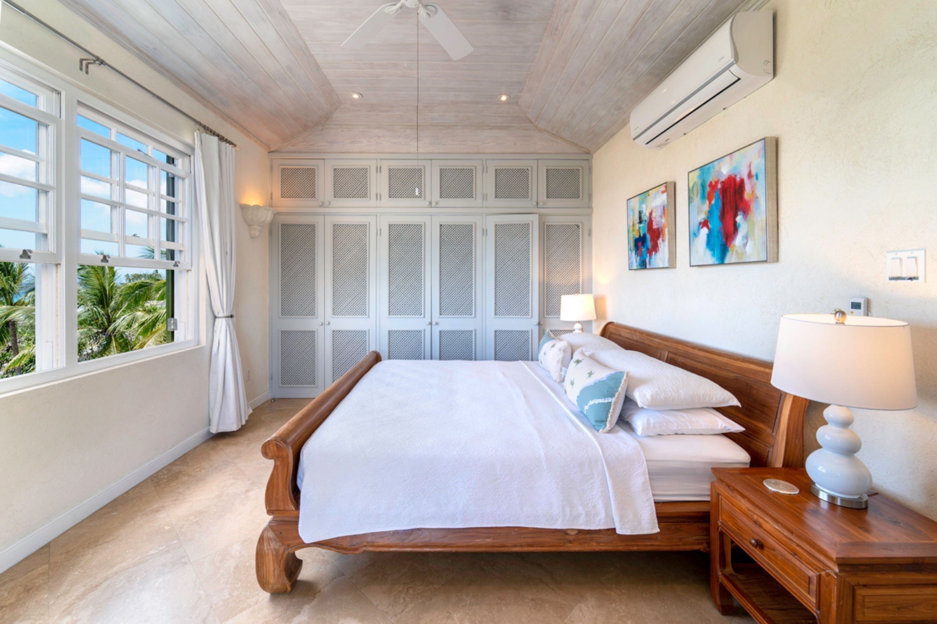 A spacious bedroom with a large bed, wooden headboard, and a nightstand with a lamp. There are multiple windows with curtains, a ceiling fan, and a wall-mounted air conditioning unit.