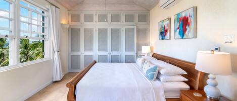 A spacious bedroom with a large bed, wooden headboard, and a nightstand with a lamp. There are multiple windows with curtains, a ceiling fan, and a wall-mounted air conditioning unit.