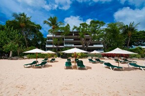 A beach with rows of green lounge chairs and white umbrellas, a multi-story building in the background, and palm trees. - Coral Cove 7 - Sunset by Blue Sky Luxury (Paynes Bay)