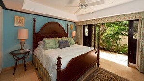 A bedroom with a wooden bed, a ceiling fan, a nightstand, and a view of greenery outside. - Coral Cove 3 - Green Flash Blue Sky Luxury (Paynes Bay)