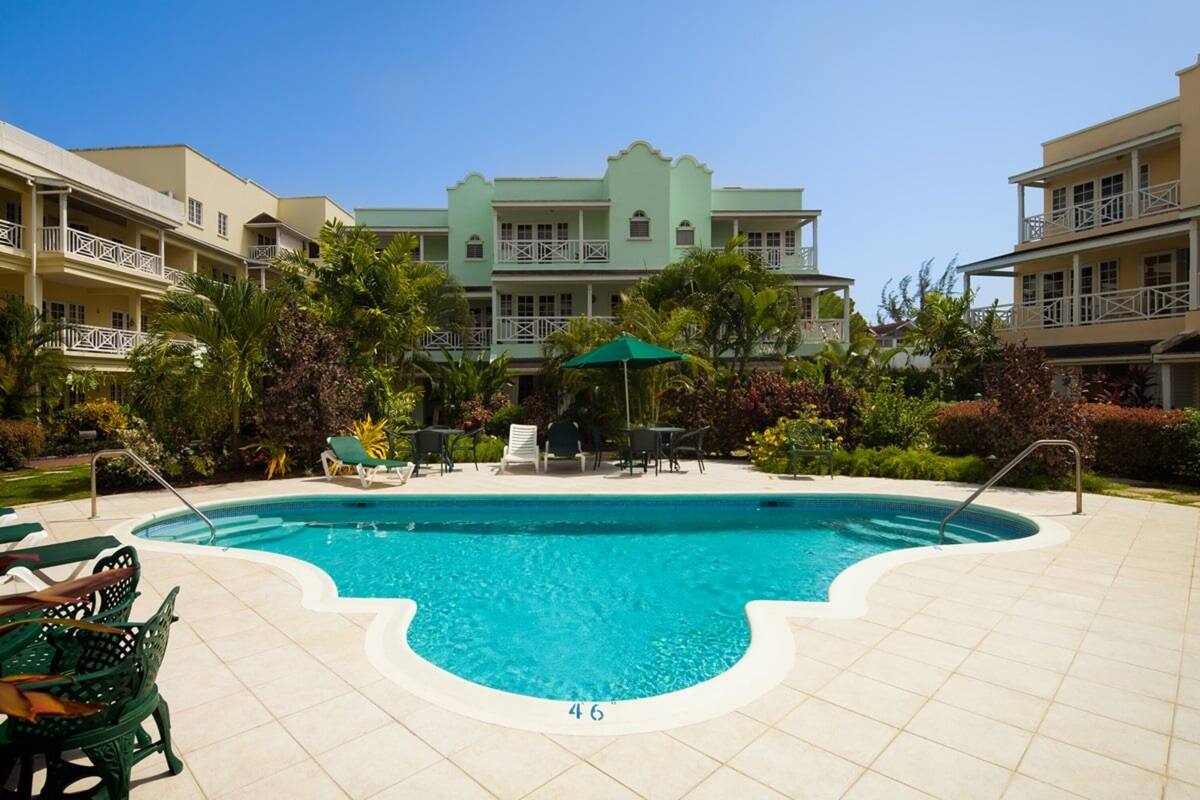 A swimming pool with a tiled deck, surrounded by lounge chairs and umbrellas, with a building in the background.