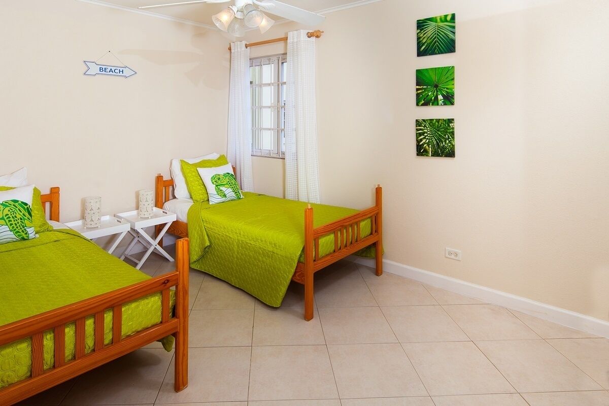 A bedroom with two beds, a window with curtains, a bedside table with a lamp, and a wall sign.