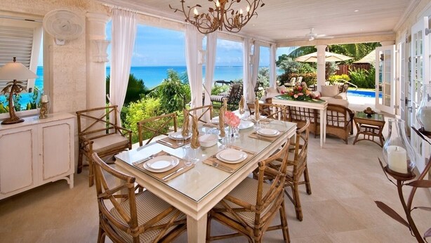 Une salle à manger spacieuse avec vue sur l’océan, comprenant une grande table dressée pour six personnes, entourée de chaises en osier.