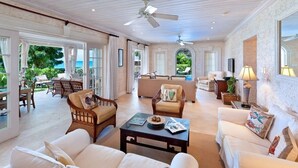 Un salon spacieux avec un grand canapé, des fauteuils et une table basse. De grandes fenêtres offrent une vue sur l’extérieur.