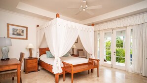 Un dormitorio con cama con dosel, ventilador de techo, escritorio con lámpara y vistas a zonas verdes a través de puertas de vidrio.