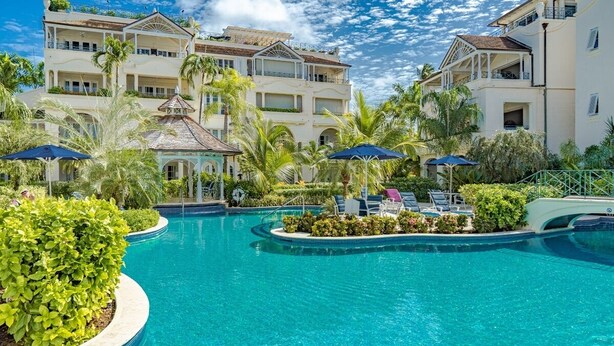 Un área de piscina con una piscina azul cristalina, sillas de descanso y un edificio con balcones y ventanas.