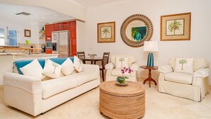 Una sala de estar con un sofá, dos sillones, una mesa de centro y una mesa de comedor. Hay cuadros enmarcados en la pared y una cocina al fondo.