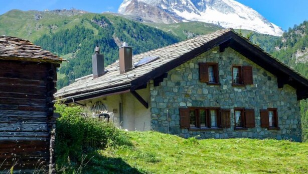 Ein Steinhaus mit geneigtem Dach und Holzhüllen, vor einer schneebedeckten Berglandschaft und grünen Hügeln gelegen.