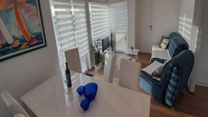 A modern living room with a dining area, large windows with blinds, a painting, and a clock on the wall.