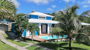 Une maison à deux étages avec une piscine, entourée de palmiers et surplombée d’un ciel bleu azur.