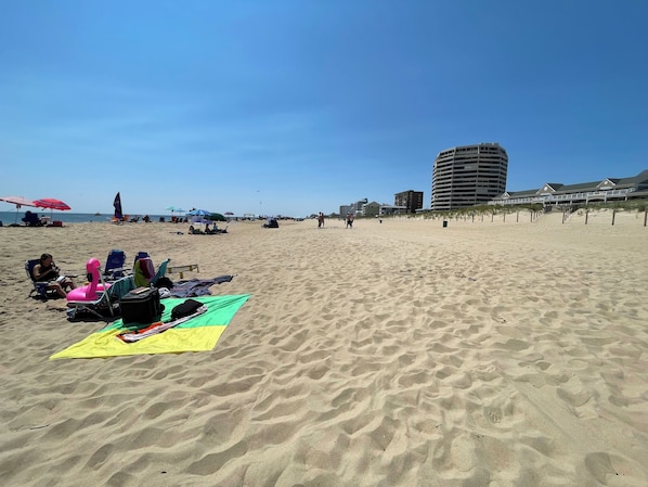 A sandy beach with beach chairs and umbrellas, a large building in the background, and a clear sky.