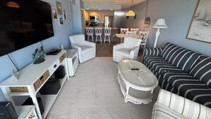 A living room with a TV, white wicker furniture, a striped sofa, and a dining area in the background. - Bimini 205 - Direct Oceanfront 87th St, WiFi, W/D, AC (Ocean City)