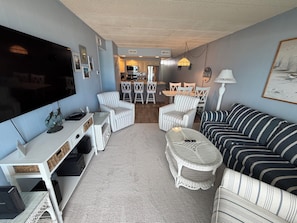 A living room with a TV, white wicker furniture, a striped sofa, and a dining area in the background.