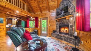 A wooden cabin interior with a stone fireplace, a leather sofa, and a dining table with a glass top. - Spacious Log Cabin with Hot Tub, Fire Pit, Large Decks & Wood Fireplace (Logan)
