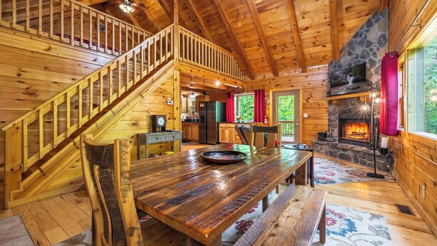 A wooden cabin interior with a staircase, dining table, benches, a stone fireplace, and a kitchen area. - Spacious Log Cabin with Hot Tub, Fire Pit, Large Decks & Wood Fireplace (Logan)