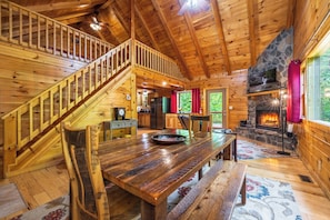 A wooden cabin interior with a staircase, dining table, benches, a stone fireplace, and a kitchen area.