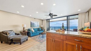 A spacious living room with a sofa, ottoman, and a coffee table. There is a kitchen area with a countertop and cabinets. - Serene Oceanfront Getaway w Pool AC - Maui Kai 304 (Lahaina)