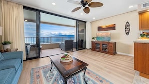 A spacious living room with a large sliding glass door leading to a balcony with a view of the ocean. - Serene Oceanfront Getaway w Pool AC - Maui Kai 304 (Lahaina)