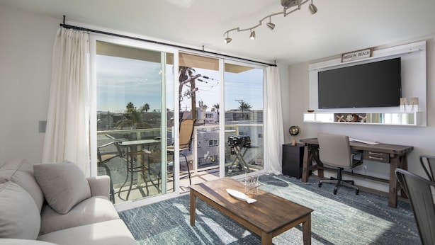 Un salon moderne comprenant un canapé, une table basse en bois, un bureau avec une chaise, un téléviseur et une porte-fenêtre coulissante donnant sur un balcon avec vue sur des palmiers et des immeubles.