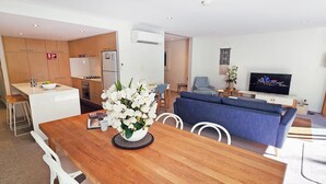 A modern apartment with a wooden dining table, a blue sofa, a kitchen with wooden cabinets, and a TV in the living area. - Stylish Parkside Retreat - Outdoor Pool & Undercover Parking - Randwick (Randwick)