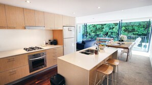 A modern kitchen with wooden cabinets, a central island with stools, a dining area with a table and chairs, and a view of greenery through large windows. - Stylish Parkside Retreat - Outdoor Pool & Undercover Parking - Randwick (Randwick)