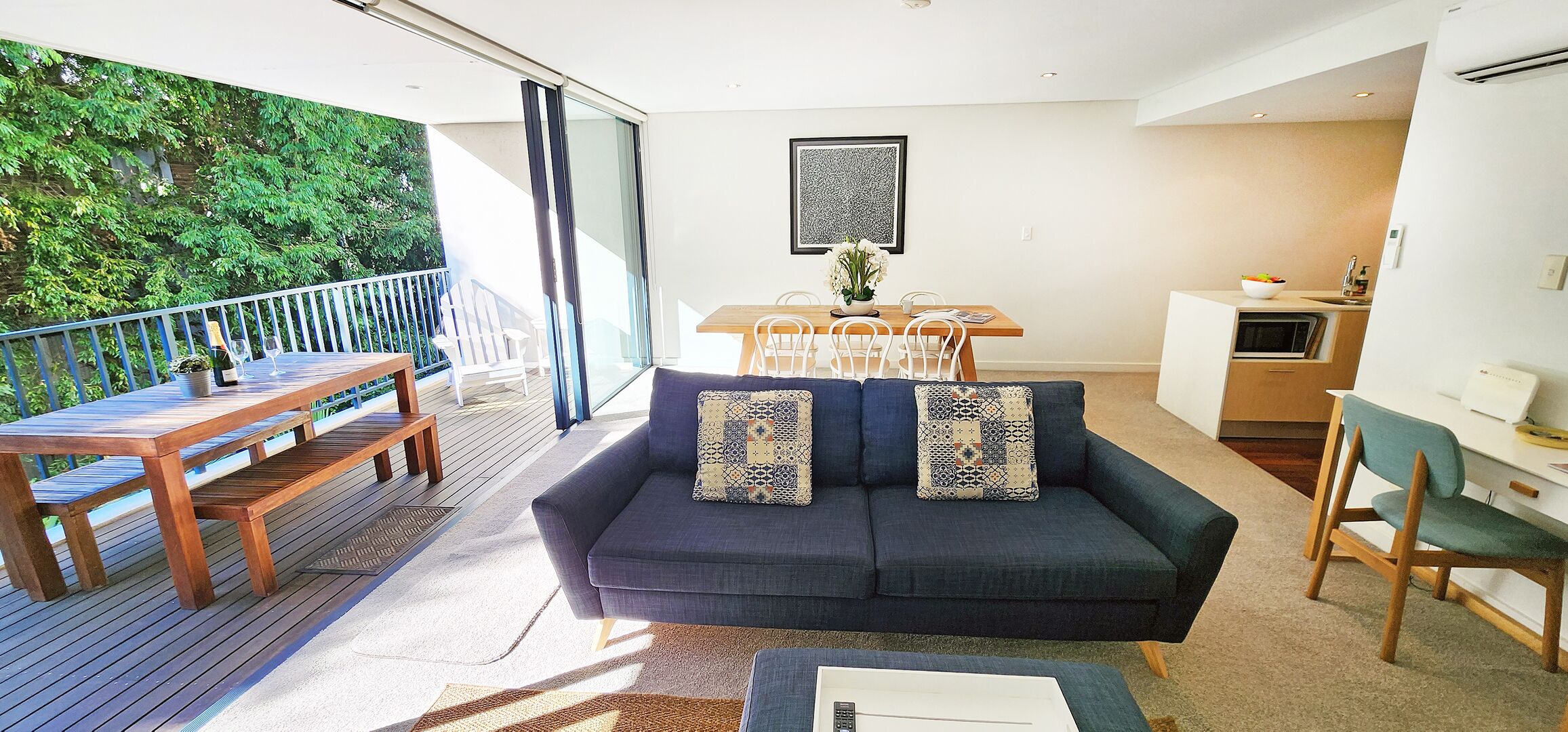 Un espacio habitable moderno con un sofá azul oscuro, mesa de comedor de madera y un balcón con vistas a zonas verdes.