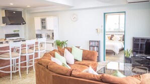 A modern living room with a brown sofa, a dining area with white chairs, a kitchen with a stove, and a bedroom with a bed and a TV. - Coco Calm (Uruma)