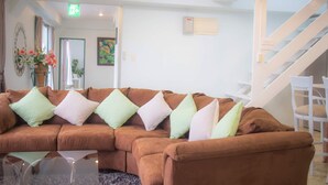 A brown sectional sofa with multiple cushions in a bright room with white walls, a staircase, and a door leading to another area. - Coco Calm (Uruma)
