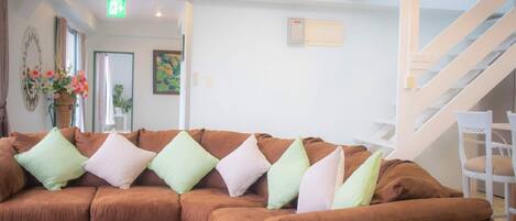 A brown sectional sofa with multiple cushions in a bright room with white walls, a staircase, and a door leading to another area.