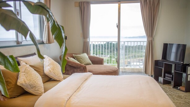 A hotel room with a large bed, a sofa, a TV, and a balcony with a view of the sea. - Coco Calm (Uruma)