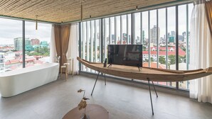 Ein modernes Zimmer mit großem Fenster, einem holzartigen, bootförmigen TV-Schrank, einem kleinen Tisch mit Vase und Blick auf die Stadt.