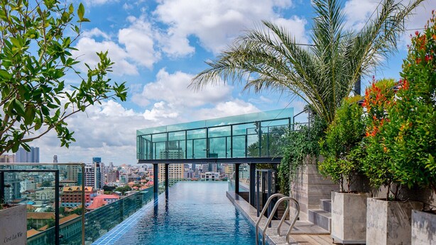 Ein Fernblick auf eine Stadtlandschaft von einem Infinity-Pool mit Glassteg, umgeben von viel Grün.