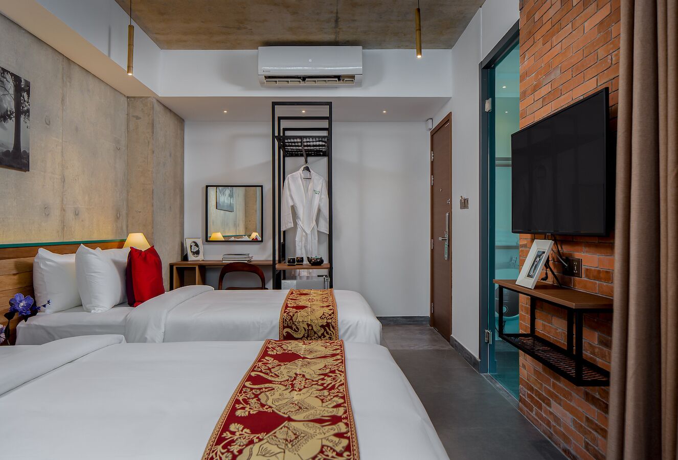 Quarto de hotel com duas camas, uma TV fixada na parede, um detalhe em parede de tijolos e uma pequena mesa com luminária.