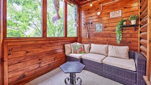 Interior acogedor de una cabaña de troncos con un sofá de mimbre, una mesita y una ventana con cortinas.
